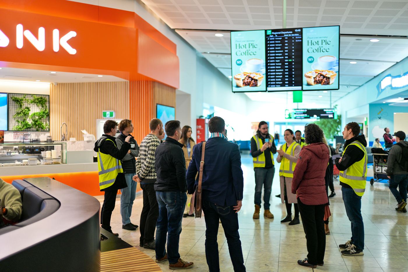 Tour group in high-visibility vests walking through the Christchurch Airport terminal food court